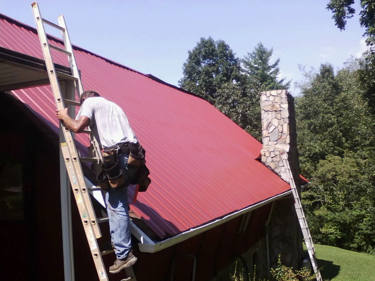 Roofer on a ladder beside a red metal roof and stone chimney — Soffit & Fascia Repair by Roofing Brothers in Cocolamus